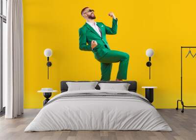 Young man celebrating success. Happy funny joyful excited guy in stylish green party suit and cool glasses raising fist up and dancing isolated on bright yellow background. Full length shot, side view Wall mural