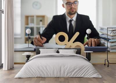 Young man credit broker or banker calculates interest rate on bank loan or income from invested investments, counting on calculator and writes result percent, sits at office desk. Selective focus Wall mural