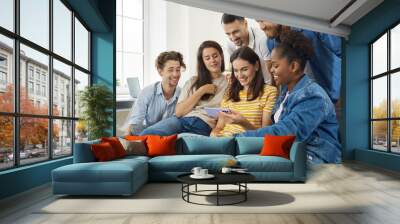 Young people watching something on a modern mobile phone. Group of happy diverse friends sitting on the sofa at home, looking at a smartphone, watching a funny video and smiling Wall mural