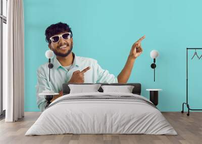 Young positive Indian man in sunglasses points with finger of two hands at copy space with smile recommends something to you, dressed in white casual shirt stands on turquoise background Wall mural