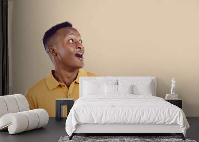 Young positive WOW African American man looks up with enthusiastic emotions when he sees unexpected promotional offer or surprise with huge discounts for shopping, stands in beige studio. Copy space Wall mural