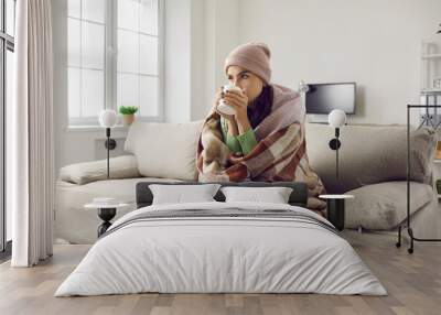 Young sick woman is treated and warmed by hot drink while sitting on sofa at home. Woman wrapped in plaid, scarf and wearing hat sitting on sofa in room with cup in hands. Home treatment concept Wall mural