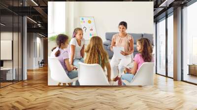 Young smiling woman psychologist conducts a mental health lesson for a group of school children girls sitting in a circle and having team work training during therapy session in classroom. Wall mural