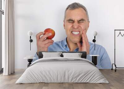 adult handsome man with grey hair wearing blue shirt touching cheek with hand suffering from sudden toothache while eating apple standing on isolated white Wall mural