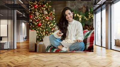 happy brunette woman wearing sweater watching tv sitting on the sofa with popcorn in room decorated for celebrating new year and christmas eve Wall mural