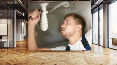 man plumber in uniform fixing the sink pipe with adjustable spanner in his hand lying on the kitchen floor looks concentrated professional plumbing repair service Wall mural