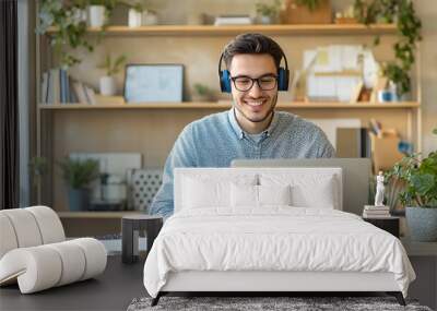 A smiling young man in headphones and glasses sits at a desk working on a laptop and making notes. Happy males in earphones watch webinars or training courses or computers and study online from home. Wall mural