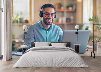 A smiling young man in headphones and glasses sits at a desk working on a laptop and making notes. Happy males in earphones watch webinars or training courses or computers and study online from home. Wall mural