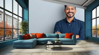 charismatic indian businessman in smart casual attire confident pose with folded hands against clean white backdrop warm smile conveys approachability and professionalism modern corporate portrait Wall mural