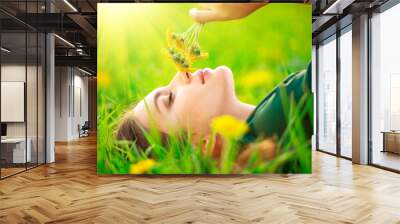 Beautiful young woman lying on the field in green grass and smelling blooming dandelions. Allergy free concept Wall mural