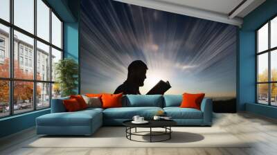 A man reading a book outdoors at sunrise, with dramatic light rays emanating from the background, symbolizing knowledge and inspiration. Wall mural