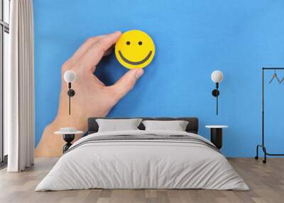 Happiness and positivity concept. Hand holding yellow smiling face in blue background with copy space. Wall mural