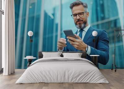 A confident businessman with a beard uses a smartphone, wearing a tailored suit, against a modern glass building backdrop. Wall mural