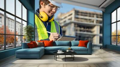 Asian male site engineer or building inspector man with green reflective vest, safety helmet, goggles and earmuffs using digital tablet inspecting working process at construction site. Wall mural