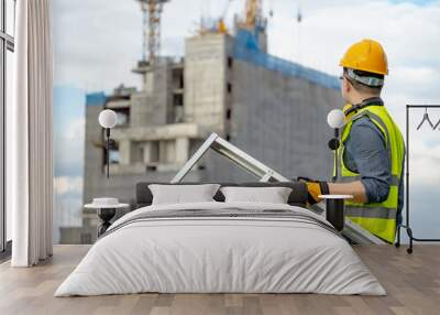 Asian man construction worker with green reflective vest, yellow safety helmet and ear muffs carrying aluminum ladder looking at unfinished building and tower crane at construction site Wall mural