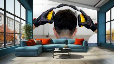 Back view of male worker with reflective orange vest and protective hand gloves pulling yellow safety ear muffs or ear protectors when wearing on his head. Equipment for high noise reduction Wall mural