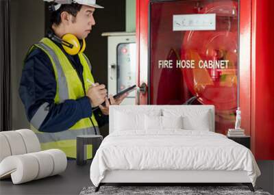 Building inspector man using digital tablet checking fire hose cabinet. Asian male worker in green vest, ear muffs and safety helmet working for building maintenance inspection Wall mural