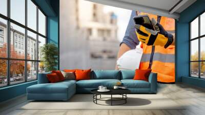 Construction worker man with orange reflective vest holding white protective safety helmet using smartphone at unfinished building site. Male engineer or foreman stay connected with social media app Wall mural