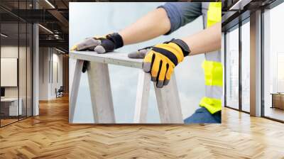 Male maintenance worker hands with protective yellow gloves holding aluminum step ladder at construction site. Building service tool and equipment Wall mural