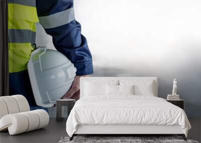 Safety workwear concept. Male hand holding white safety helmet or hard hat. Construction worker man in protective suit and reflective green vest standing with building concrete floor in the background Wall mural