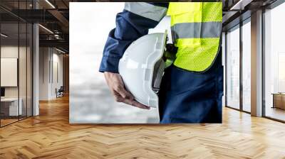 Safety workwear concept. Male hand holding white safety helmet or hard hat. Construction worker man in protective suit and reflective green vest standing with building concrete floor in the background Wall mural