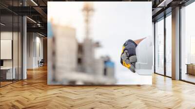 Safety workwear concept. Male hand holding white safety helmet or hard hat. Construction worker man with reflective orange vest and protective gloves standing at unfinished building with tower crane Wall mural