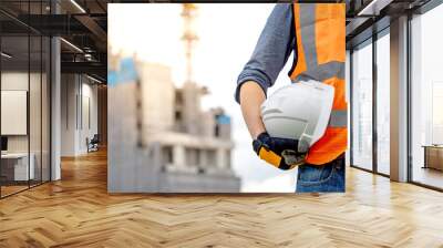 Safety workwear concept. Male hand holding white safety helmet or hard hat. Construction worker man with reflective orange vest and protective gloves standing at unfinished building with tower crane Wall mural