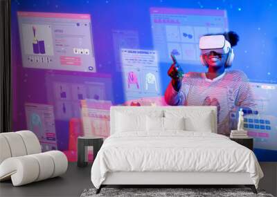 African girl looking list while wearing VR glass and sitting at sofa with shopping bag. Teenager using augmented reality glasses while choosing and buying product at neon color background. Ingenuity. Wall mural