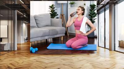 Athletic and sporty woman drinking water on fitness mat after finishing home body workout exercise session for fit physique and healthy sport lifestyle at home. Gaiety home exercise workout training. Wall mural