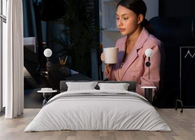 Beautiful sitting businesswoman with pink suit drinking coffee while searching business project report data, waiting email back with accepted win bidding job on laptop at late night time. Postulate. Wall mural