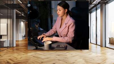 Beautiful sitting businesswoman with pink suit working on business project report data on laptop, analyzing and calculating of financial data overworked at late night time at modern office. Postulate. Wall mural