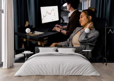 Businesswoman working on desk with stretching arm up and down manner with body health ache of strain overwork while coworker trading stock market on website on pc at late over night time. Infobahn. Wall mural