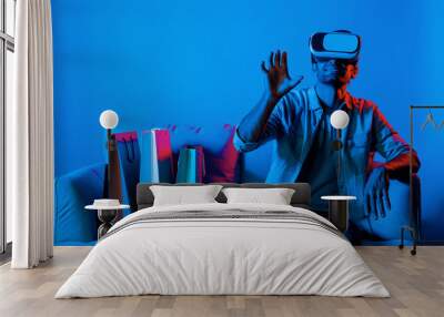 Caucasian man with vr glass posing pointing finger while sitting at sofa with shopping bags. Person selecting and doing shopping online by using VR technology and virtual goggle at home. Deviation. Wall mural