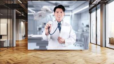 Energetic doctor holding document while standing at operation room. Asian healthcare worker wearing lab coat and stethoscope while working at hospital and posing at camera. Healthcare. Ingenuity. Wall mural