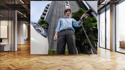 Fisheye view suit-clad progressive businessman at charging station for electric vehicle at modern city with the residential apartment condo building in the background. Idea for eco-friendly car. Wall mural