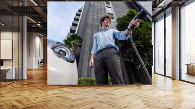 Fisheye view suit-clad progressive businessman at charging station for electric vehicle at modern city with the residential apartment condo building in the background. Idea for eco-friendly car. Wall mural