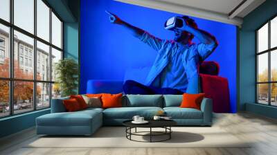 Man pointing and choosing program in simulated metaverse by using VR glasses. Person with headset sitting at sofa while entering in virtual world by using futuristic technology equipment. Deviation. Wall mural