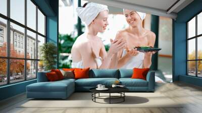 Portrait of two attractive woman in towel giggling during hold the herbal bowl surrounded by natural spa environment. Pretty girls with beautiful skin using herbal scrub at spa salon.Tranquility. Wall mural