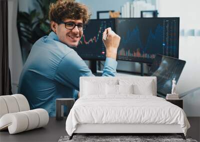 Profile of smiling young stock trader with curly hair looking at camera, sitting against on financial exchange screen at modern office. Concept of dynamic business digital investment. Gusher. Wall mural