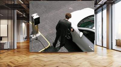 Progressive businessman unplugs charger plug from charging station to his electric car before driving around city center. Eco friendly rechargeable car powered by sustainable and clean energy. Wall mural