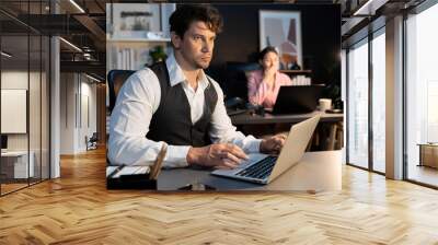 Sitting man with serious face thinking creative business project planning at night lighting time at modern office with blurry secretary checking paper report with folder background concept. Postulate. Wall mural