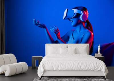 Smart female sitting on sofa surrounded by neon light wear VR headset connect metaverse, futuristic cyberspace community technology, watch and interacting with virtual object with hand. Hallucination. Wall mural