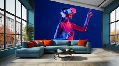 Smart female stand with surrounded by cyberpunk neon light wear VR headset connecting metaverse, futuristic cyberspace community technology. Woman using finger pointing virtual object. Hallucination. Wall mural