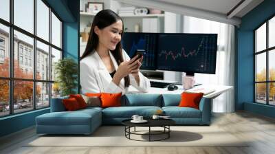 Smiling young Asian businesswoman with happy face looking on smartphone in profitable of exchange stock market graph at modern office. Concept of investing high profit in wealth security. Stratagem. Wall mural