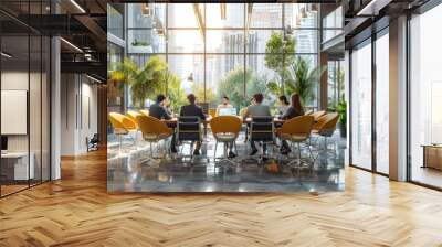 The photo shows a group of people sitting around a table in a modern office space. There are plants and large windows in the background. The people are all wearing business casual attire and are Wall mural