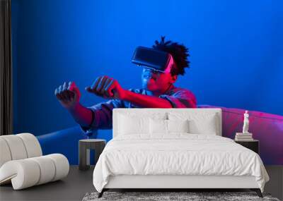 Young African American player looking VR turning drive car racing gaming meta on pink blue neon metaverse world digital futuristic technology virtual reality at living room sitting sofa. Contrivance. Wall mural