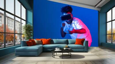 Young African American player using VR turn gun shooting hunter 3D gamer in hologram blue pink neon lighting wall connecting digital futuristic technology virtual reality metaverse world. Contrivance. Wall mural