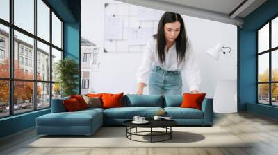 Young beautiful caucasian architect puts blueprint on the table and compare with house model while inspect hos model construction carefully at modern office with blueprint striped behind. Immaculate. Wall mural