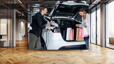 Young couple travel with EV electric car to shopping center parking lot charging in downtown city showing urban sustainability lifestyle by green clean rechargeable energy of electric vehicle innards Wall mural