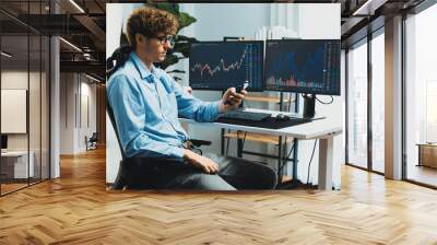 Young investor stock trader looking on smartphone, analyzing dynamic financial exchange investment on two laptop screens background in current market graph in real time at modern office. Gusher. Wall mural